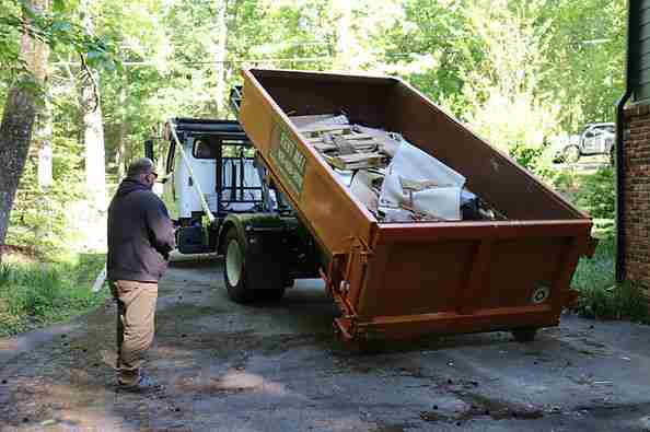 Operator beside a tilted orange dumpster containing debris on a roll-off truck